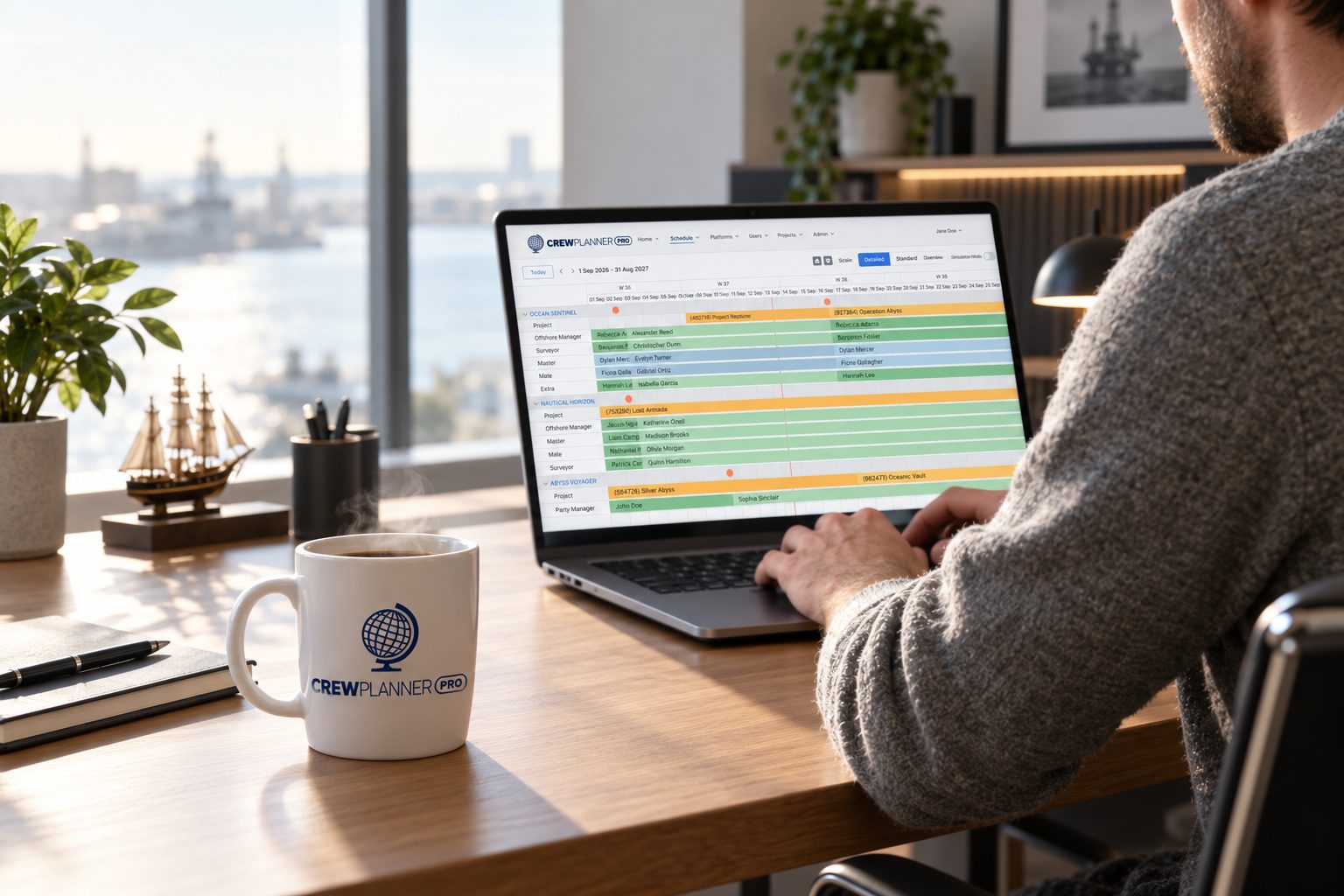 A crewing coordinator working at a desk with a laptop showing Crew Planner Pro, coffee mug branded with the Crew Planner Pro logo beside the keyboard.